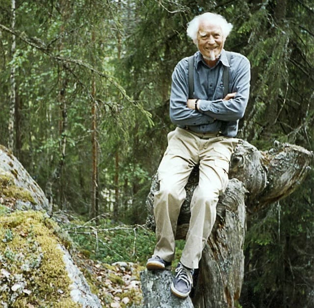 Åke Forsberg, vithårig och leende med armarna i kors, sittande på en gammal trädstam i skogen.
